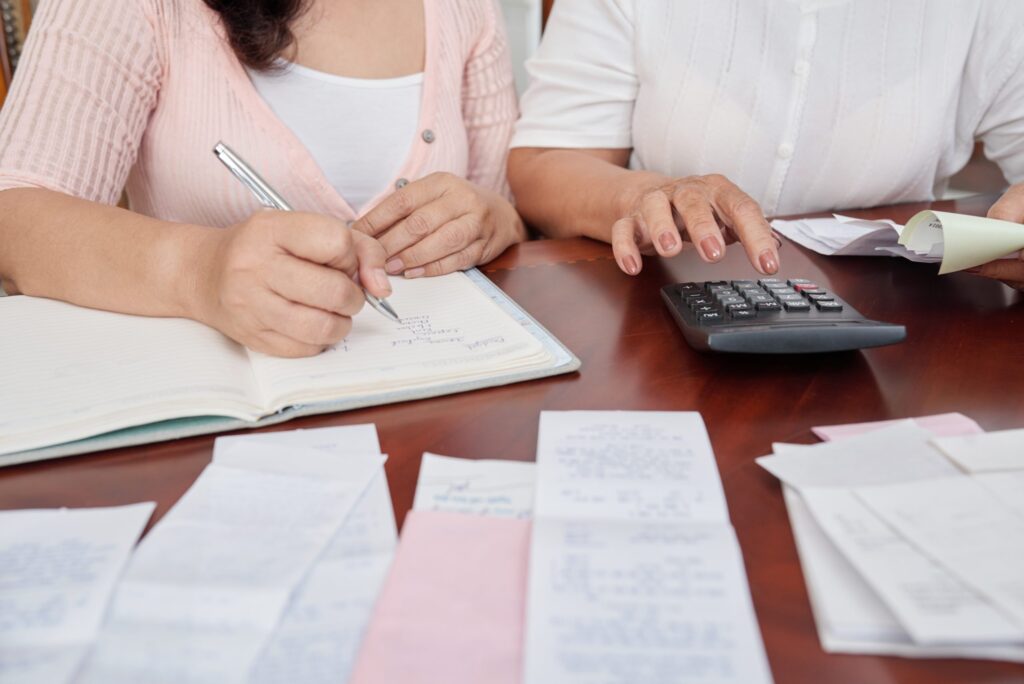 Mulheres sentadas à mesa com recibos, usando calculadora e anotando notas no bloco de notas. Todos os dados referentes a viagens corporativas